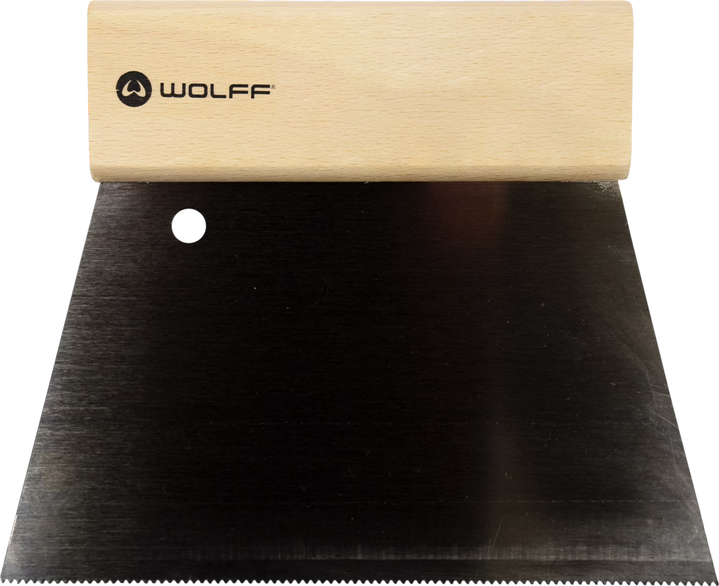 Close-up of a wide metal putty knife or drywall taping knife with a light wooden handle stamped with the black 'WOLFF' logo. The tool features a broad, flat black steel blade with a round hanging hole near the top and a finely serrated edge along the bottom. The wooden handle shows a smooth grain and rounded shape for comfortable grip. The blade displays a subtle brushed texture and minor scuffs; the photo is taken from above with the handle at the top and the long blade extending downward against a white background. The implement is typically used for spreading and smoothing plaster, putty or adhesive and for scraping surfaces.
